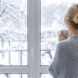 Woman holding a cup of coffee looking out the window at her snowy backyard.