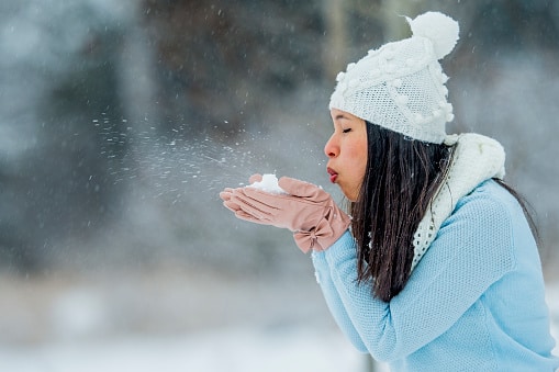 A woman blows snow from her cupped hands on a snowy winter day.