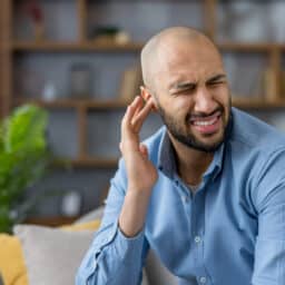 Young man feeling intense ear pain, touching his ear with a grimace, experiencing discomfort and health problems needing treatment for otitis or hearing issues